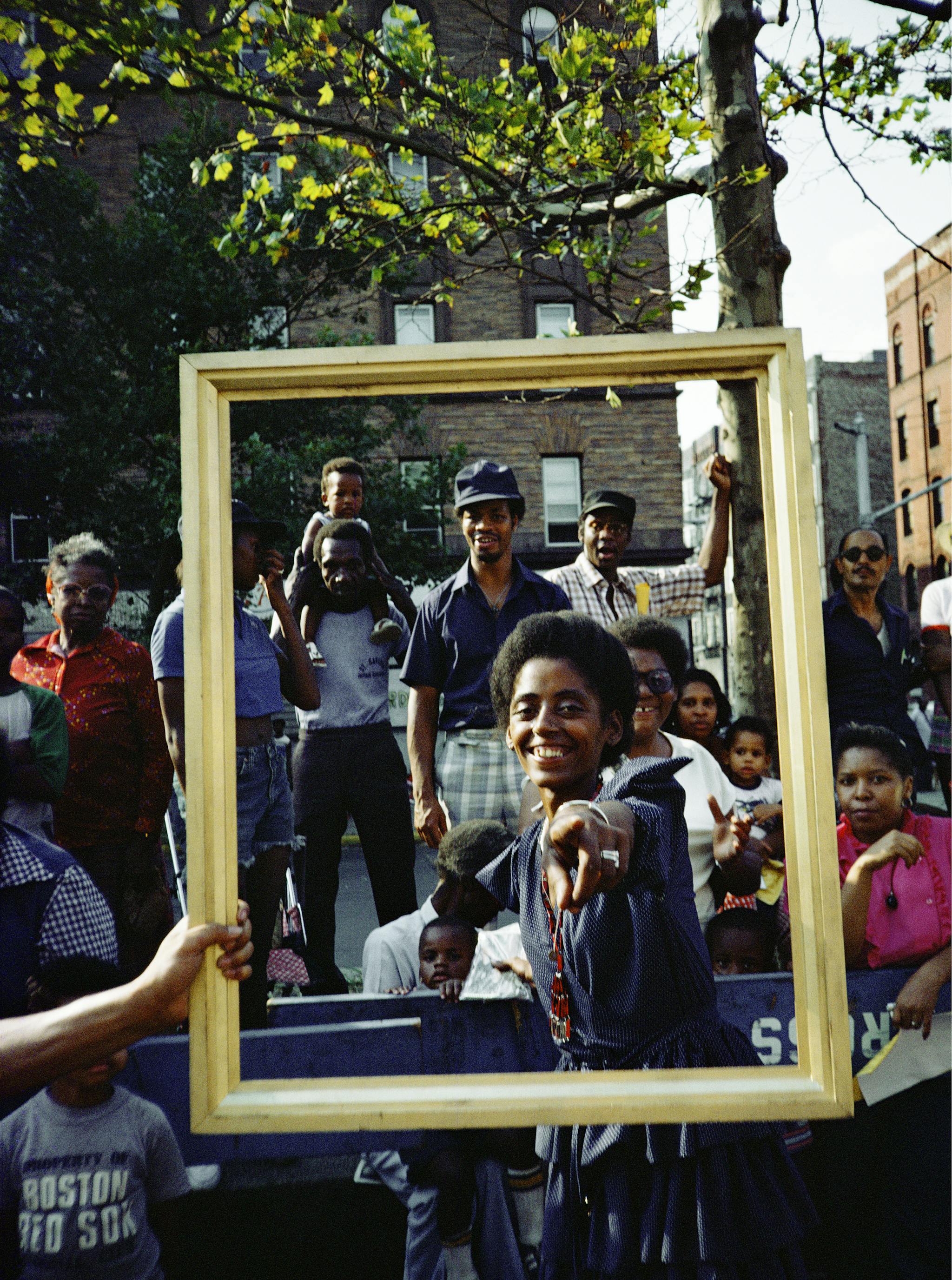 A group of figures appear within a wooden picture frame, while a young person points directly at the camera, smiling.