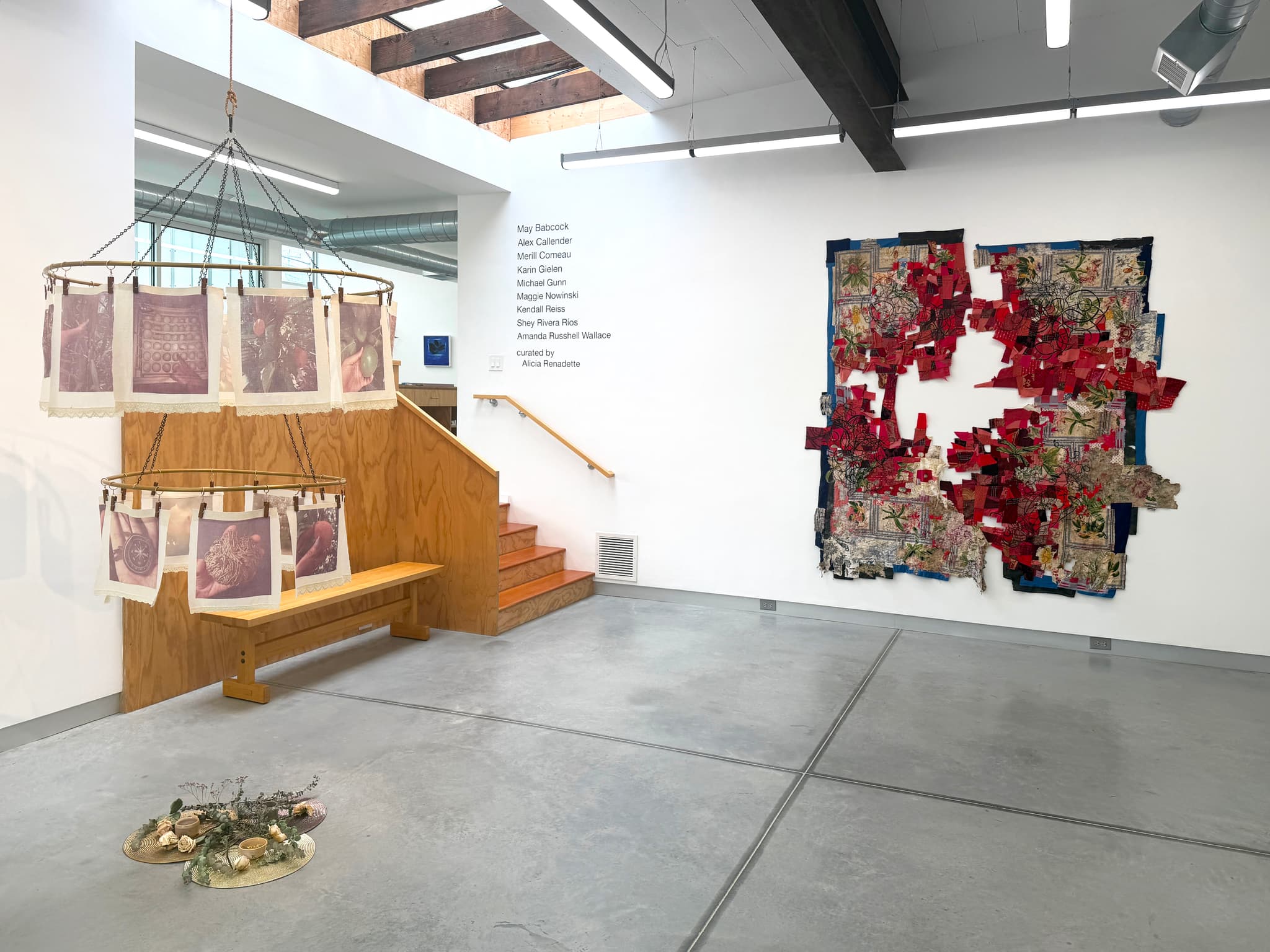 A two-tier sculpture hangs to the left of a large, textile collage on a gallery wall.