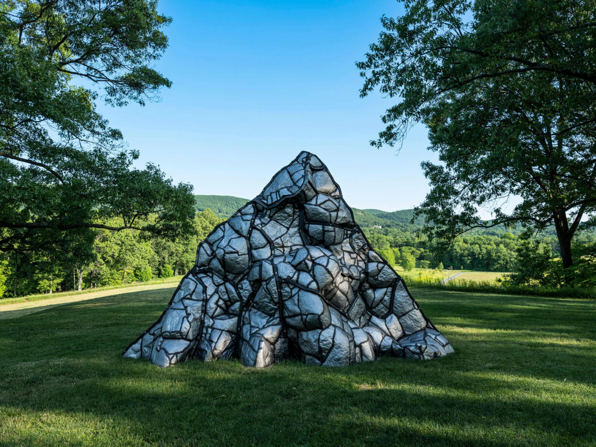 A public art installation representing a volcano stands in a grassy, green park area, between trees.