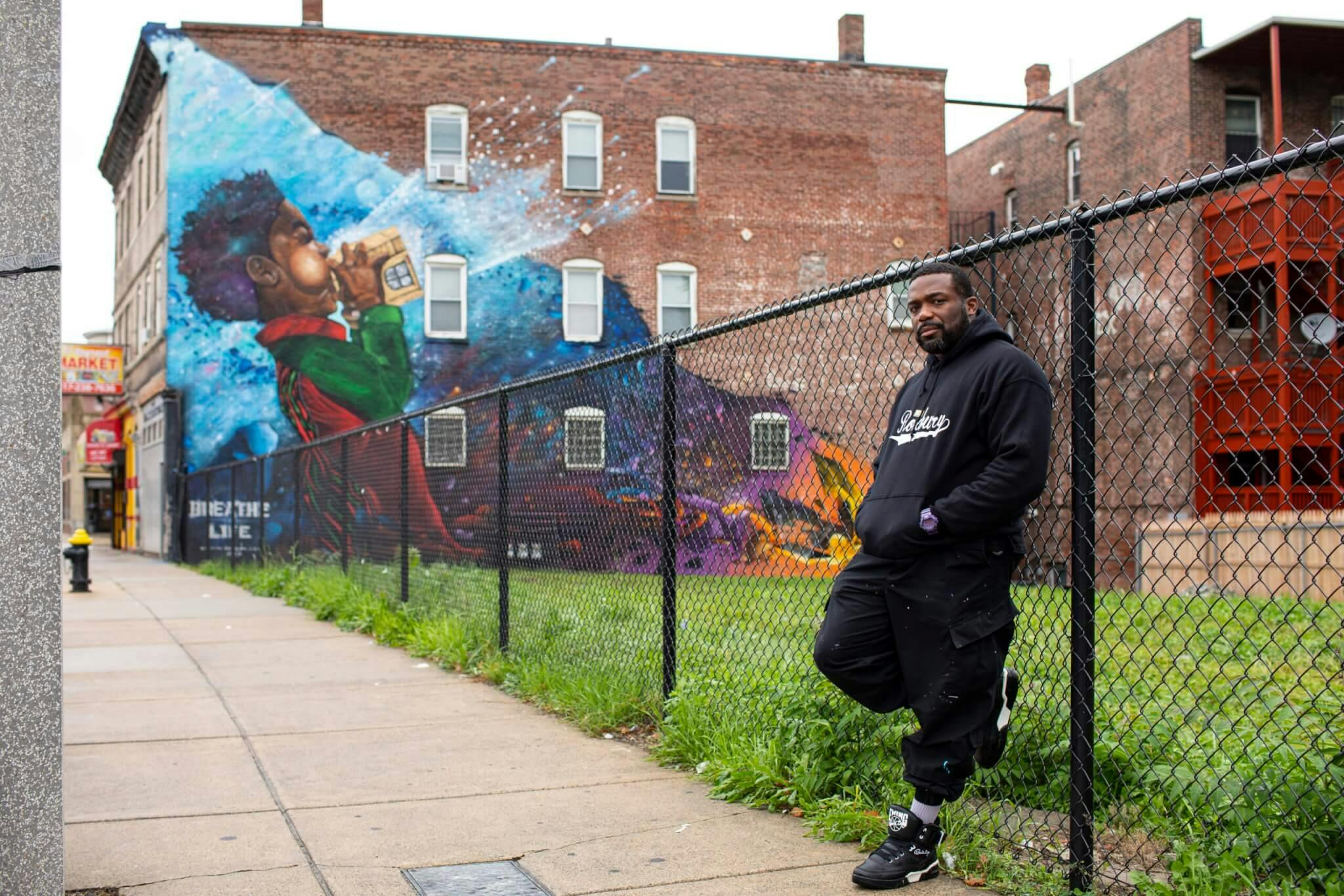 An artist stands by a fence in front of a colorful mural, in the neighborhood of Dorchester.