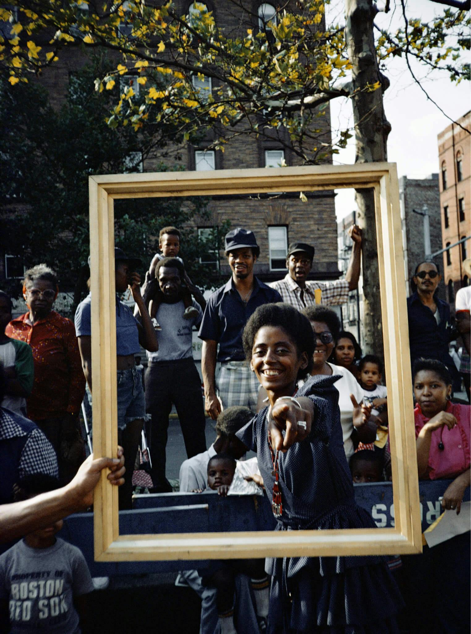A group of figures appear within a wooden picture frame, while a young person points directly at the camera, smiling.