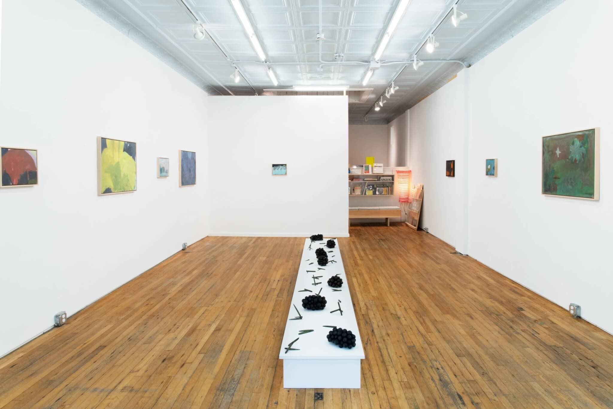 Sculptures lie on a narrow, white table; paintings line the walls of a gallery.