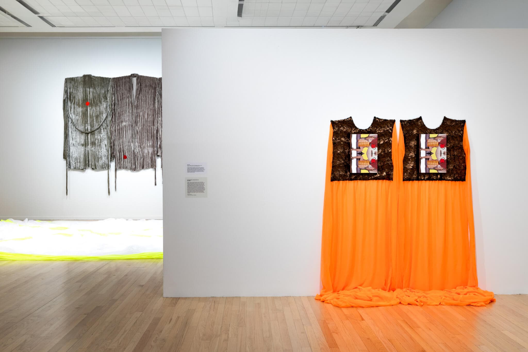 Two orange, flowing dresses hang from the wall of a gallery space.