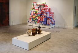 A sculpture made up of colorful cardboard bricks rests on a pedestal against a wall; small brick-based sculptures are presented on a pedestal at the center of the white-walled room.