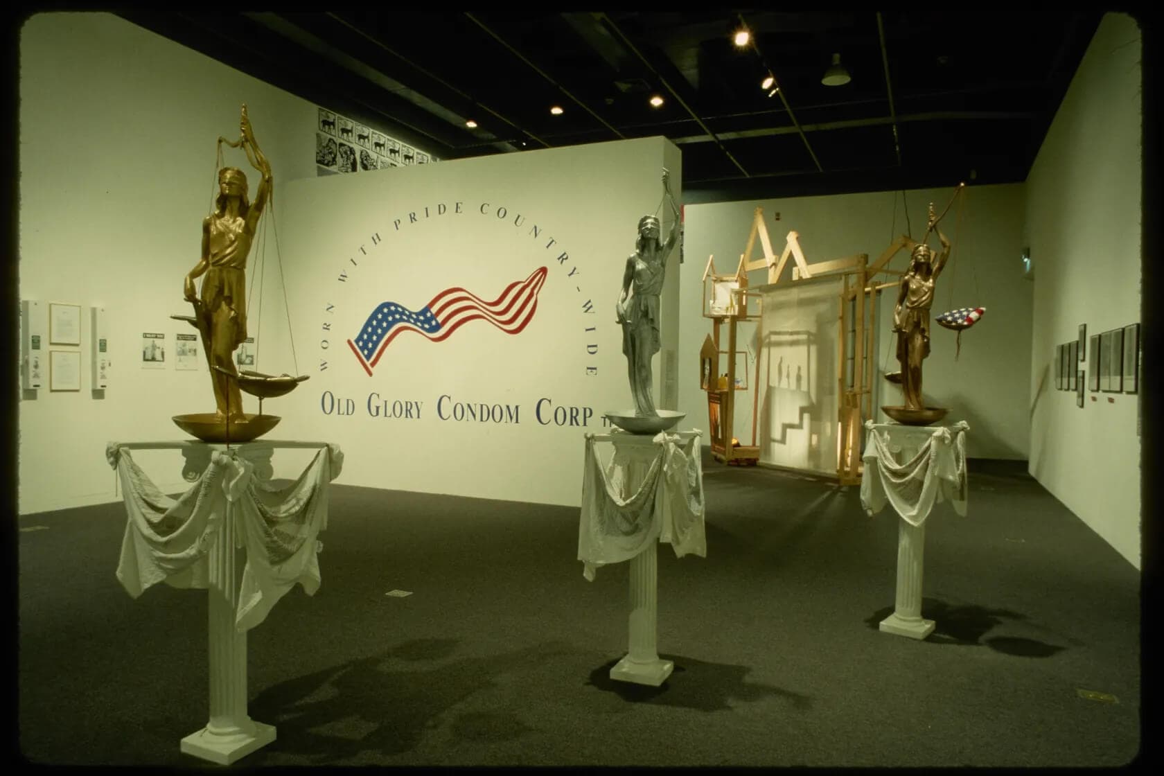 An installation view. A white wall with the Old Glory Condom Corp logo sits behind three statues.
