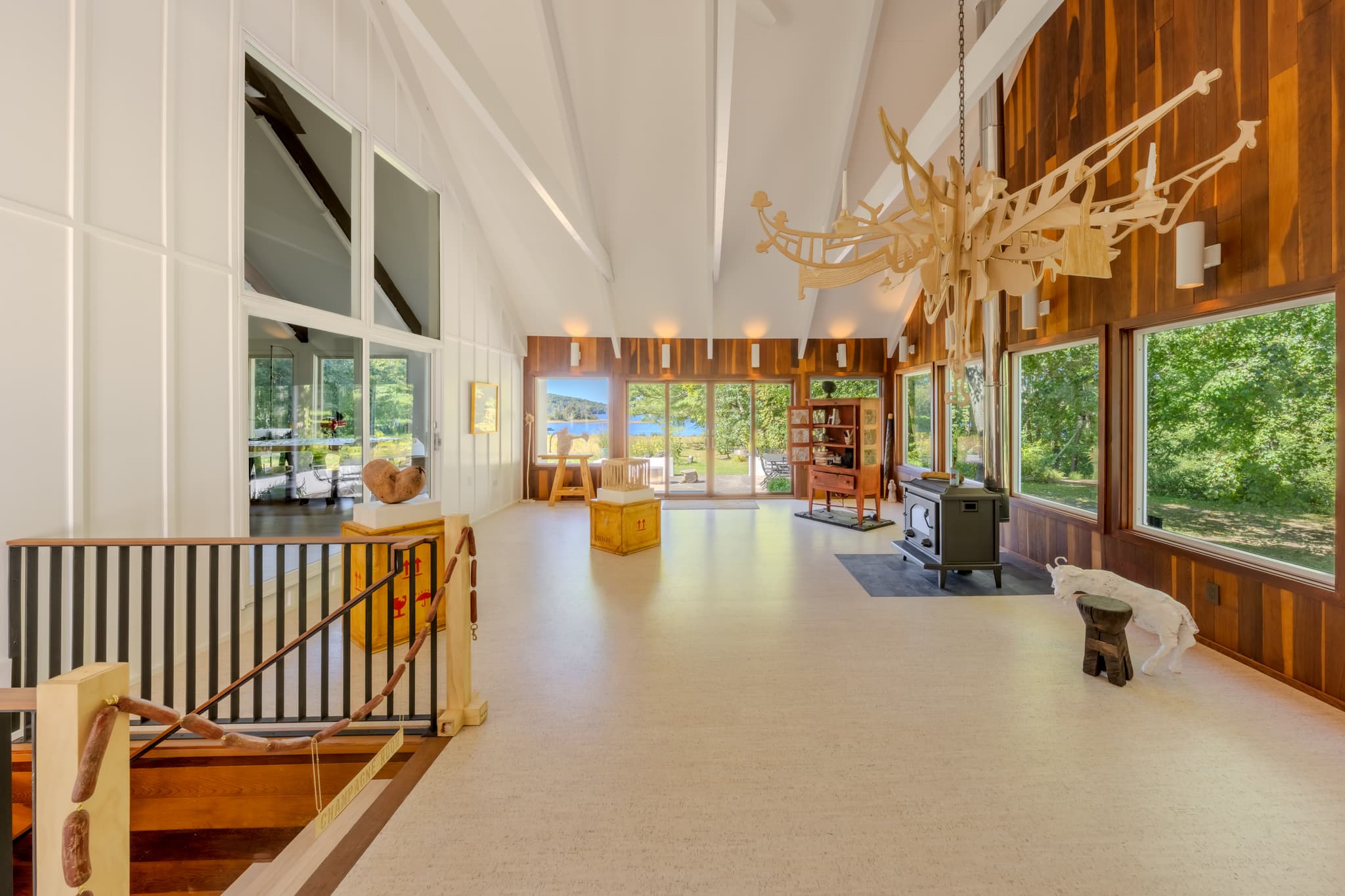 A large wooden chandelier hangs in the foreground above smaller sculptures situated around a window-lined room, with views of nature beyond.